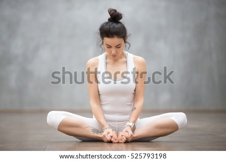 Portrait of beautiful young woman with floral tattoos working out against grey wall, doing yoga or pilates exercise, sitting in baddha konasana, bound angle, cobbler, butterfly pose. Full length shot Royalty-Free Stock Photo #525793198
