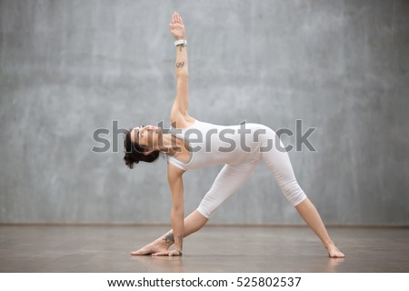 Profile portrait of beautiful young woman wearing white sportswear working out against grey wall, doing yoga or pilates exercise. Standing in Utthita Trikonasana, extended triangle pose. Full length Royalty-Free Stock Photo #525802537