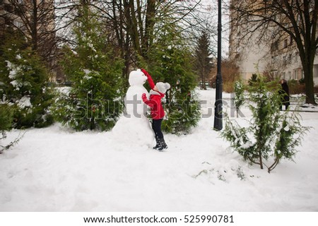 The girl of school age builds a snowman. On the ground there is a lot of snow. The girl in a red jacket and a gray knitted cap stands near a big snowman. She decorates the head of a snowman.