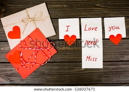 Red envelopes with ropes and paper hearts lie behind white cards with lettering 'I love/need/miss you' on wooden table