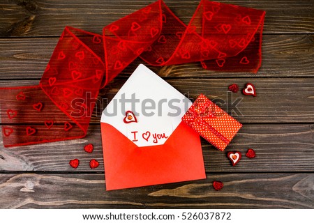 Red envelope with card 'I love you' inside lies among red decorative hearts on wooden table