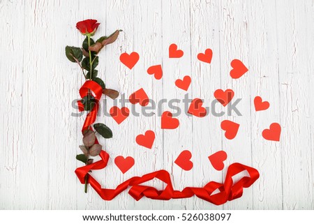Red rose with long red silk ribbon surrounded with paper hearts lies on white wooden floor