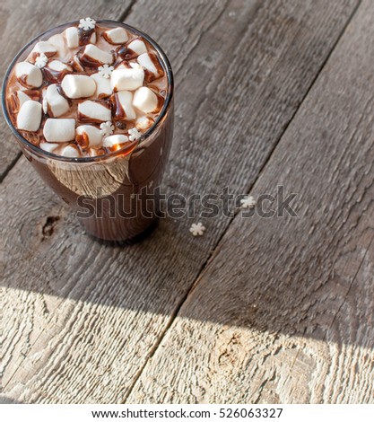 Winter hot cocoa with marshmallows, chocolate syrup, sugar snowflakes in a tall glass on a wooden background.