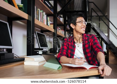 Picture of happy asian male student dressed in shirt in a cage and wearing glasses using tablet at the library and writing in notebook. Look aside and smiling.
