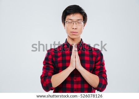 Picture of serious young man dressed in casual shirt in a cage and wearing eyeglasses make praying gesture and isolated over white background