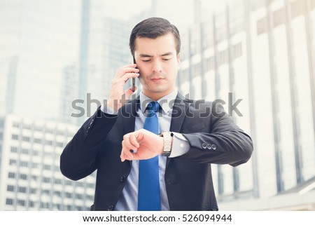 Businessman looking at his watch checking time while calling on cell phone in blur office building background