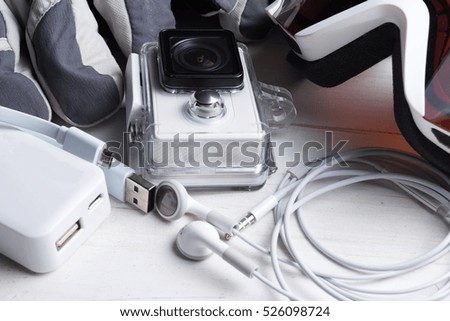 White technology on natural wooden table background. Power bank, action camera, head phones, cable goggles, gloves. Active winter leisure time sports holidays abstract concept. Skiing, snowboarding.