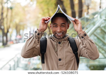 Image of young happy african man with notebook on his head outdoors. Looking at camera.
