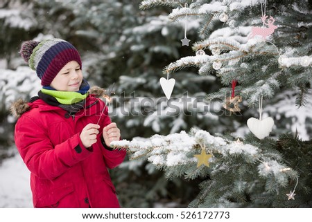 Happy smiling kid boy decorating Christmas tree in winter snow park. Child having fun. Christmas celebration outdoors. Winter and holidays concept.
