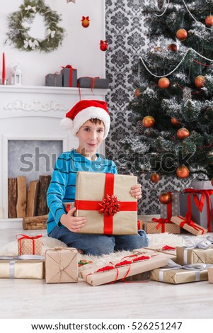 Cute happy boy in santa hat unwrap christmas present box on holiday morning in beautiful room interior. Male child open Xmas gifts near big decorated fir tree and fireplace. Winter holidays concept