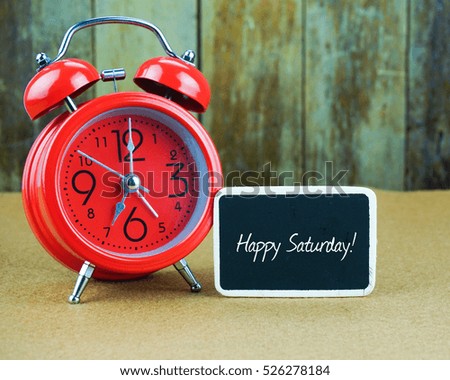 HAPPY SATURDAY! inscription written on chalkboard, red alarm clock on desk with old wooden background. Time concept.