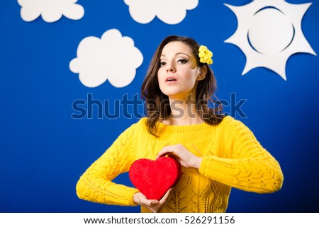 Happy pretty girl in yellow sweater with red heart shaped present box, portrait over bluescreen background with paper clouds and sun