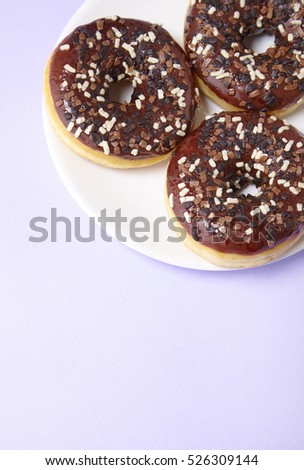 A plate full of iced chocolate sprinkle ring donuts on a pastel purple background with blank space below
