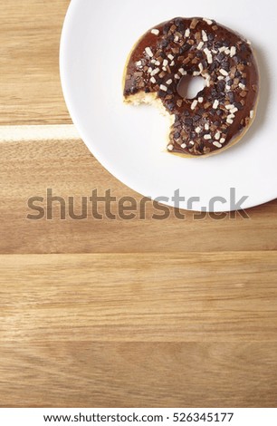 An iced chocolate sprinkle ring donut with bite mark, on a wooden kitchen counter background with empty space below