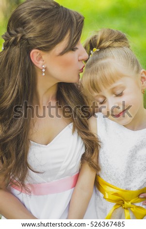 beautiful mother and cute little daughter spend together a weekend in nature, relaxing, mother tenderly kissing the head daughter, dressed in white dresses
