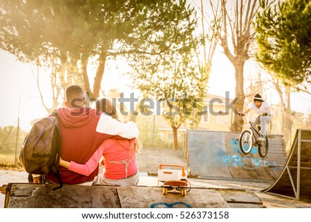 Multiracial couple in love sitting at skate park with music watching friends on bmx freestyle exhibition - Urban relationship concept with young people having fun outdoors - Warm contrasted filter
