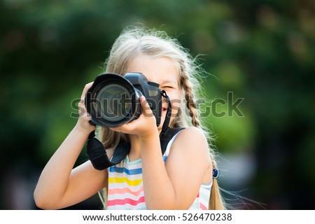 Portrait of cheerful smiling little girl making photo with big camera in hands outdoors
