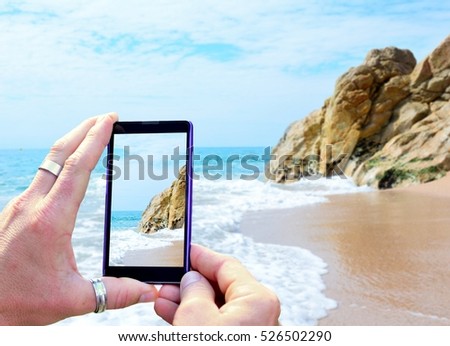 View over the mobile phone display during taking a picture of Costa Brava beach in Calella, Spain. Holding the mobile phone in hands and taking a photo. Focused on mobile phone screen. 