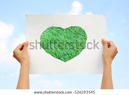 Hands holding paper art sheet Burning in shape of heart with image of green grass against Cloud sky.