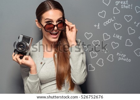 Happy photographer gained likes and hearts in social networks depicted on the grey wall with a chalk. Hipster lowers her glasses with one hand and holds a retro camera.