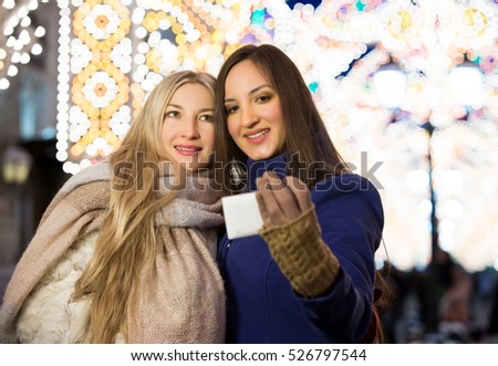Blond and brunette sisters using smart phone taking pic on a night street with Christmas light decorations. Friendship, love and travel concept. Girls making selfie.