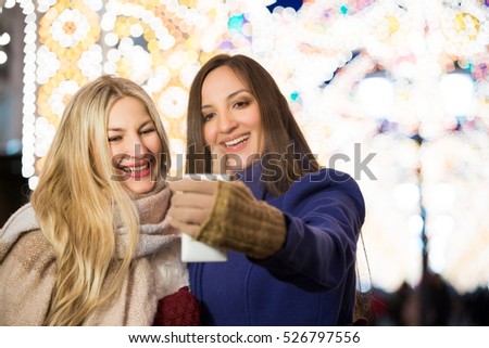 Blond and brunette sisters using smart phone taking pic on a night street with Christmas light decorations. Friendship, love and travel concept. Girls making selfie.