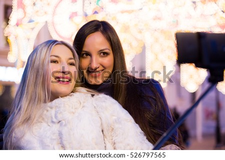 Blond and brunette sisters using selfie stick taking pic on on a night street with Christmas light decorations. Friendship, love and travel concept. Girls in trend