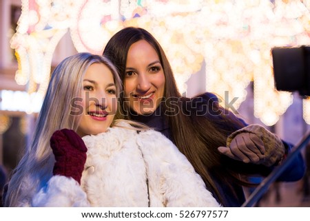 Blond and brunette sisters using selfie stick taking pic on on a night street with Christmas light decorations. Friendship, love and travel concept. Girls in trend