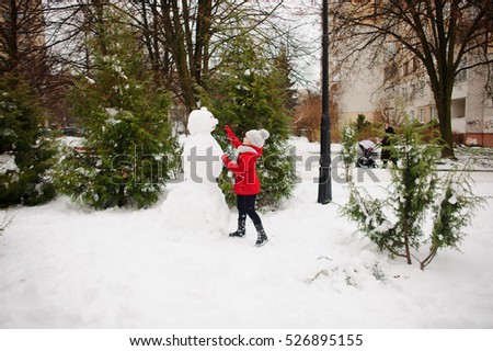 The girl of school age builds a snowman. On the ground there is a lot of snow. The girl in a red jacket and a gray knitted cap stands near a big snowman. She decorates the head of a snowman.