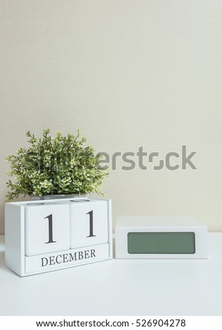 White wooden calendar with black 11 december word with clock and plant on white wood desk and cream wallpaper textured background , selective focus at the calendar
