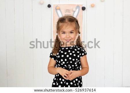 Smiling little girl in black and white dress and the picture of bunny behind her back placed in such way that hare'??s ears look like her own ones, happy childhood concept, indoor studio portrait 