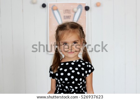 Smiling little girl in black and white dress and the picture of bunny behind her back placed in such way that hare'??s ears look like her own ones, happy childhood concept, indoor studio portrait 