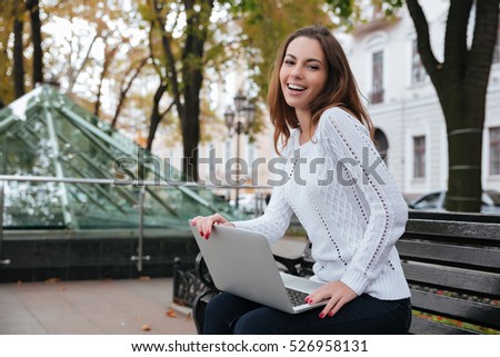 Happy attractive young woman using laptop on bench in park
