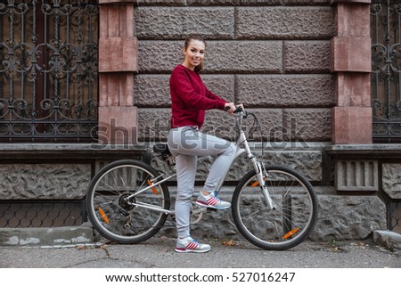 Picture of cheerful woman dressed in sweater walking with her bicycle in the city while standing near wall and smiling. Look at camera.