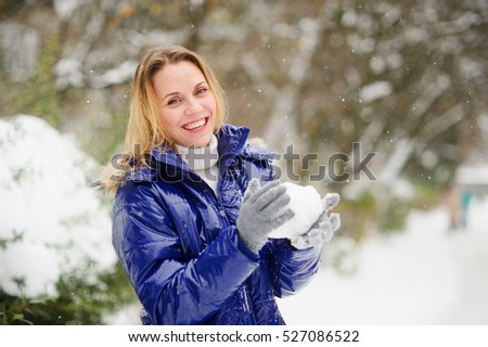 Beautiful young woman plays snowballs. It is snowing. Earth is covered with snowdrifts. Woman is dressed in a bright blue down-padded coat, without headdress. Excited by play, she does not feel cold.