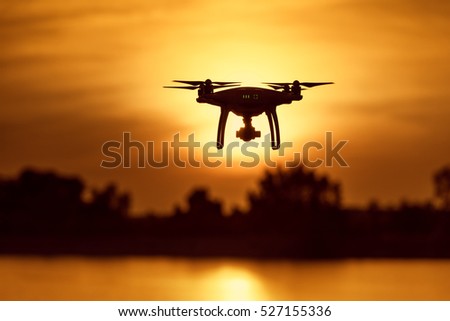 Silhouette of modern RC Drone / Quadcopter with camera flying above the river with tree background at sunset
