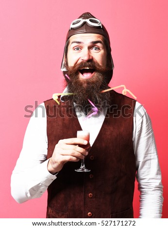 handsome bearded pilot or aviator man with long beard and mustache on smiling face holding glass of alcoholic shot in vintage suede leather waistcoat with hat and glasses on red studio background