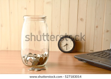 The coin inside of bottle ,alarm clock and lap top on the wood table.