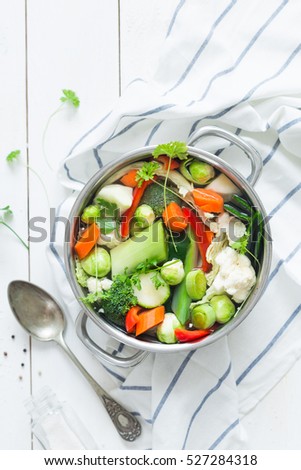 Various fresh vegetables in a pot - colorful fresh clear spring soup (vegetarian bouillon or stock). Rural kitchen scenery from above (top view). White rustic wooden background.