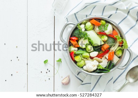 Various fresh vegetables in a pot - colorful fresh clear spring soup (vegetarian bouillon or stock). Rural kitchen scenery from above (top view). White wooden background - layout with free text space.