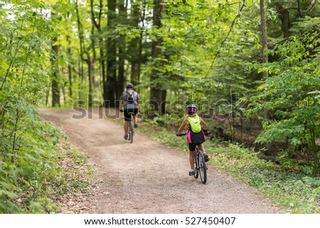 Couple of people cycling together in the woods down a dirt path on a beautiful summer day. Green canopy of trees above them. View from the back of people - full body. Royalty-Free Stock Photo #527450407