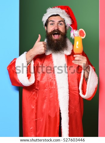 handsome bearded santa claus man with long beard on smiling face holding glass of nonalcoholic cocktail in christmas or xmas sweater and new year hat showing cool on colorful studio background