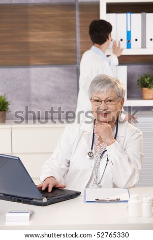 Smiling doctor sitting at desk with laptop, assistant looking at folder in background.