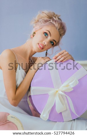 portrait of a luxurious blonde in a white dress sitting on a chair with a round gift box in hand on a lilac background