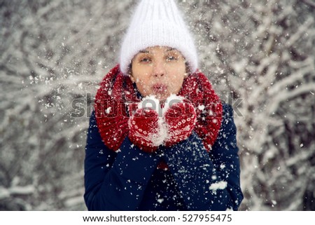 young girl in white knitted hat, red scarf and mittens blows snow with hands in snowy woods