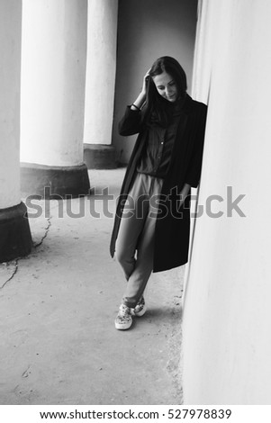 Young and fashionable business woman wearing long coat staying by the wall and columns of ancient building in black and white style