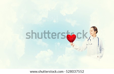 Young woman doctor against sky background holding red heart