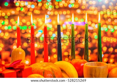 Kwanzaa celebration concept with seven candles red, black and green, gift box, pumpkin, bowl and fruit on light blur bokeh background, close up