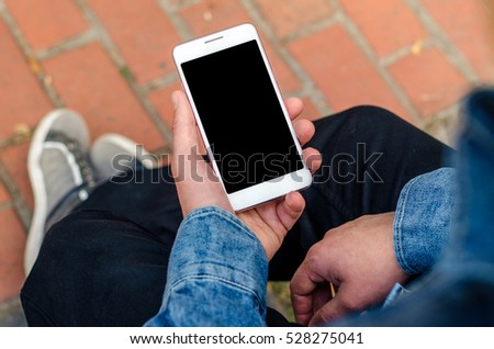close up white mobile phone in hands a young hipster trendy dressed business man on a background of a stone paving slabs and red brick. Business man typing on mobile and look mobile banking. Royalty-Free Stock Photo #528275041