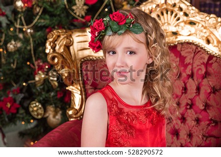 Beautiful blonde in a long red dress at the Christmas tree. Portrait of a girl with floral wreath on her head. Beauty sitting in a chair